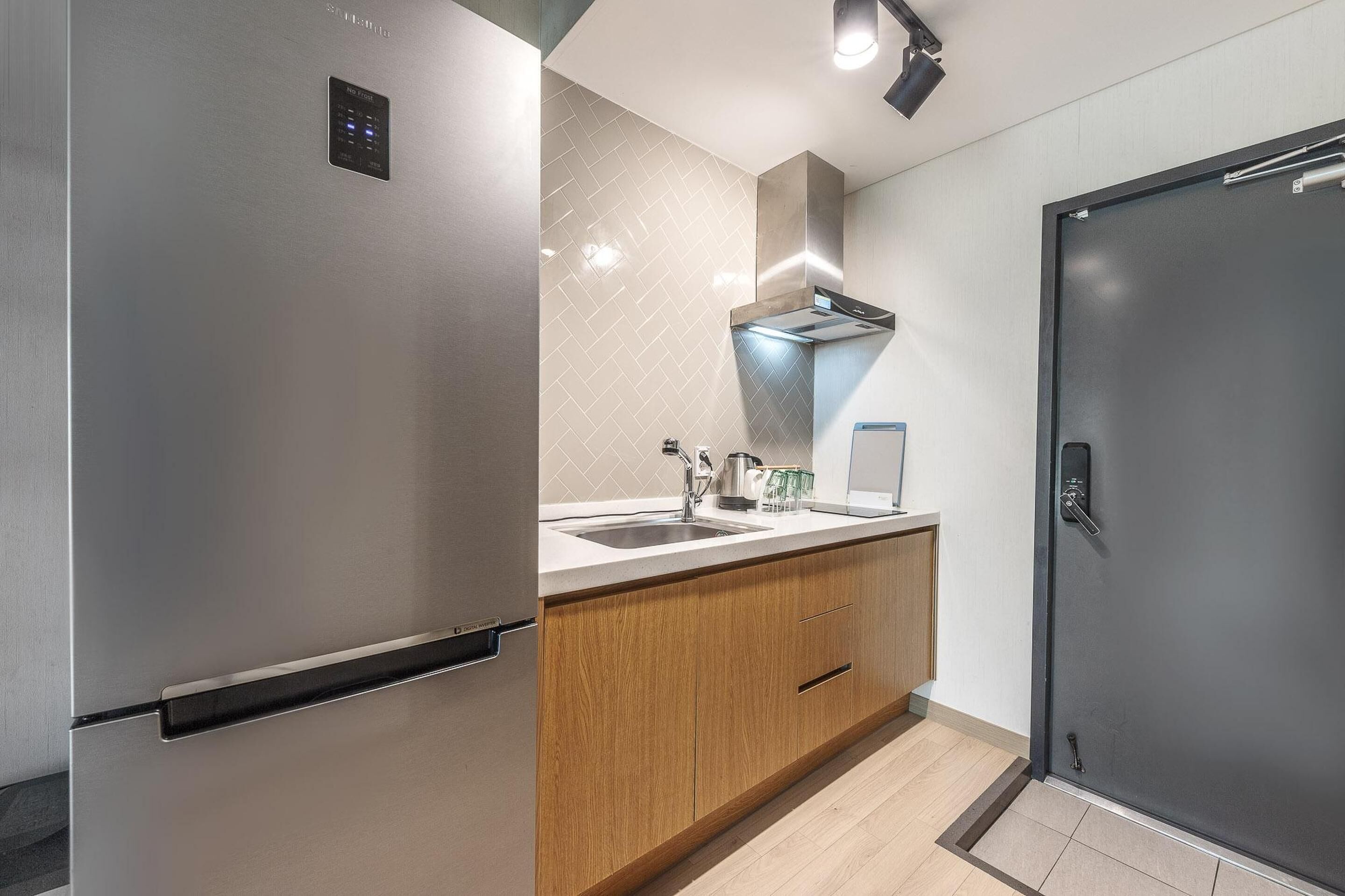 A modern kitchen with a stainless steel refrigerator, wooden cabinets, a built-in oven, and a black door.