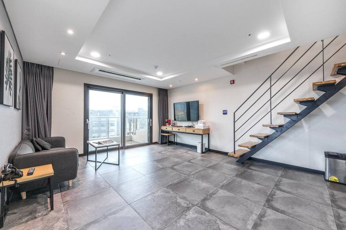 A modern hotel room with a large window, a sofa, a desk, and a staircase.