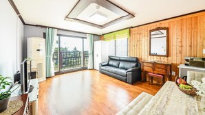 Un salón amplio con un sofá de cuero negro, suelo de madera, un ventanal con cortinas y una pared de madera con un espejo, junto a una pequeña mesa con una cesta de frutas.