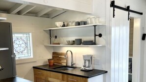Une cuisine moderne avec des meubles blancs, un plan de travail noir et une machine à café. On y trouve une porte coulissante de style rustique et une fenêtre ornée d’un motif géométrique.