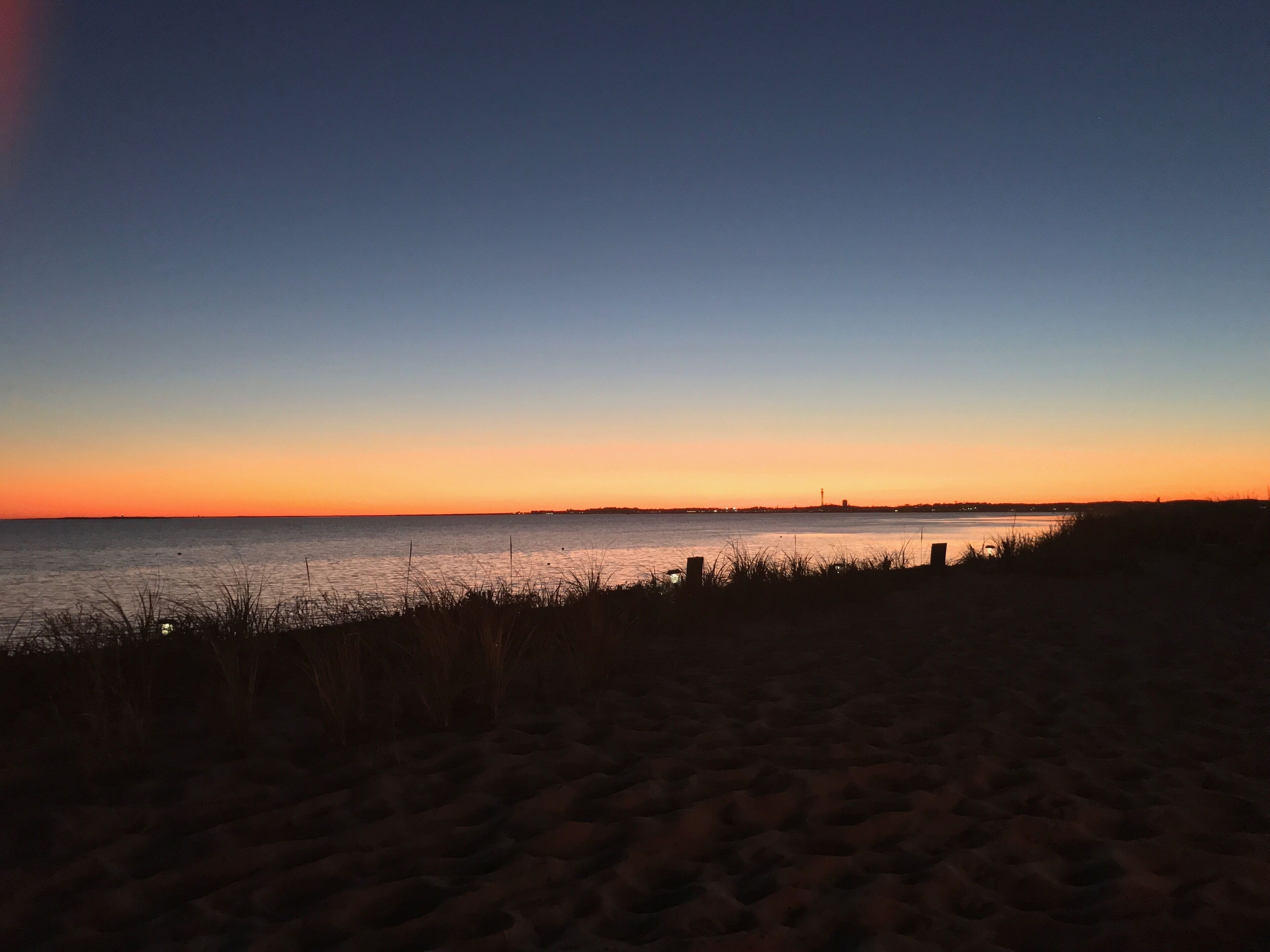 Una playa al atardecer con cielo despejado, agua tranquila y silueta de la ciudad a lo lejos.