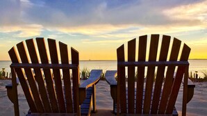Deux fauteuils Adirondack en bois, placés face à une plage, avec un coucher de soleil en arrière-plan.