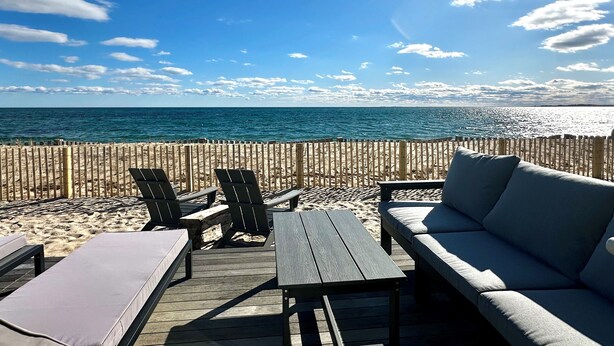 Un espace de détente aménagé au bord de la plage, avec une terrasse en bois, des chaises longues et un canapé, offrant une vue sur l’océan.