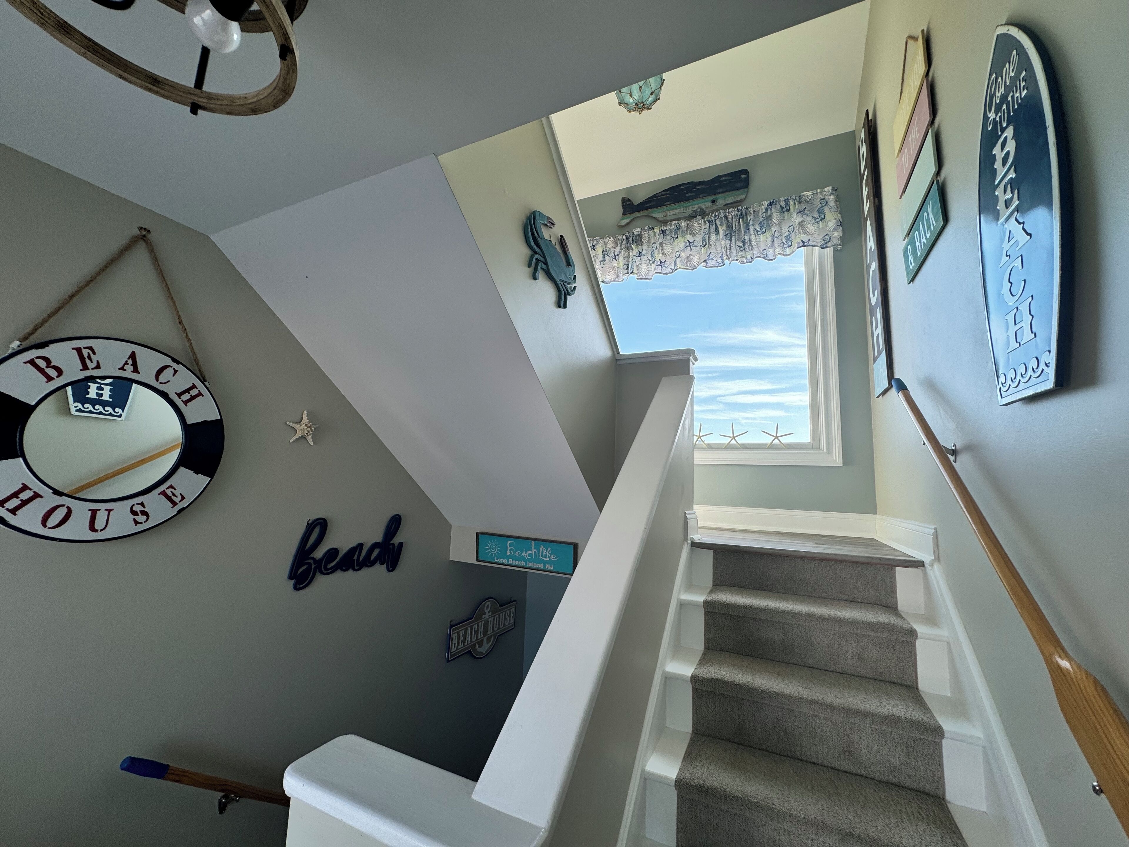 A staircase with a view of the beach through a window, decorated with nautical-themed wall art including a 'Beach House' sign and starfish.