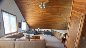 A loft-style bedroom with a sloped ceiling, a wooden ceiling fan, a large bed, a wooden railing, and a view of the outdoors through a window.