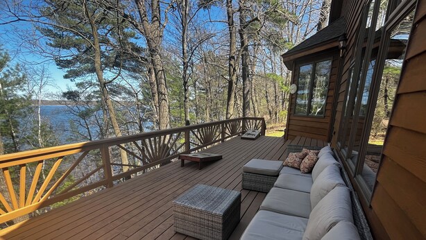 A wooden deck with a sofa and wicker ottoman, overlooking a forested area and a body of water.