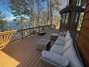 A wooden deck with a sofa and wicker ottoman, overlooking a forested area and a body of water. - Moose Lodge on Big Yellow Lake (Webster)