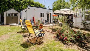 Un cortile con un piccolo capanno, una zona patio con tavolo e sedie, e un giardino fiorito.