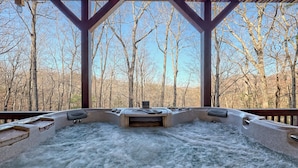 A hot tub with a stone exterior, surrounded by a wooden deck and a metal railing, overlooking a forested area. - Ultra Peaceful Retreat! Large Cabin, 2 Masters on Main, Games & Lake! (Ellijay)