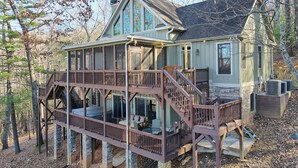 A two-story house with a wooden deck and a stone foundation, surrounded by trees. - Ultra Peaceful Retreat! Large Cabin, 2 Masters on Main, Games & Lake! (Ellijay)