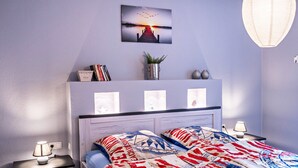 Ein Schlafzimmer mit einem Bett, Nachttischen, einem Wandregal mit Büchern und einer Pflanze sowie einem Wandbild.