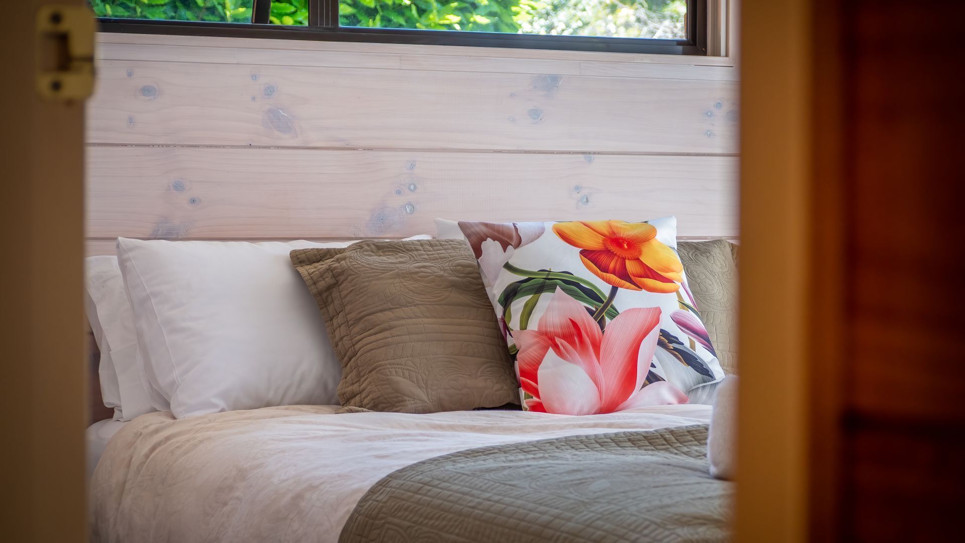 A bed with white and beige bedding, a floral pillow, and a window with a view of greenery.
