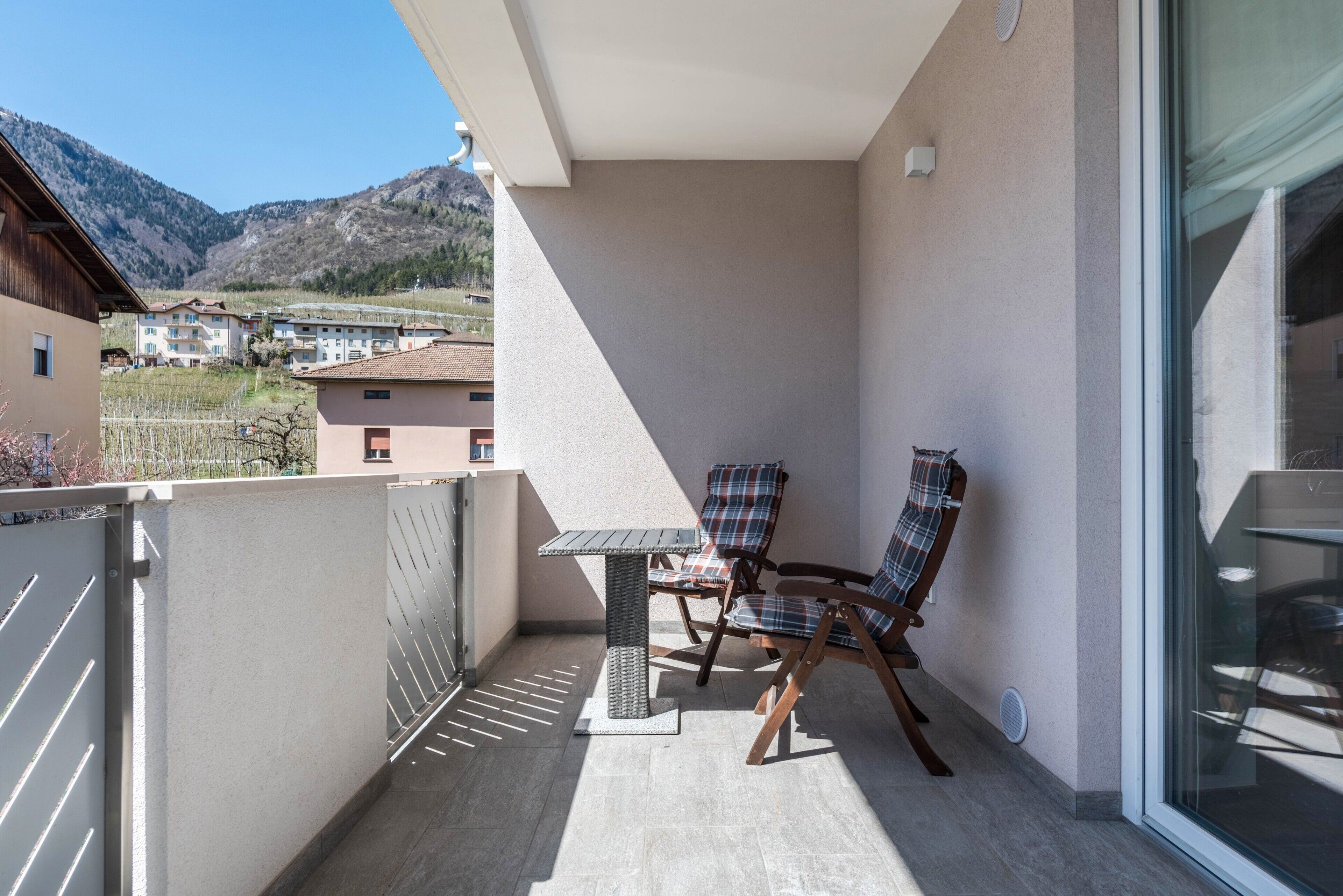 Un balcon avec une table et deux chaises, une vue sur des bâtiments et des montagnes en arrière-plan, et une porte vitrée donnant sur un espace intérieur.