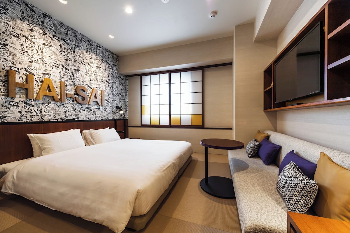 A modern hotel room with a large bed, a sofa, a small round table, and a TV mounted on the wall.
