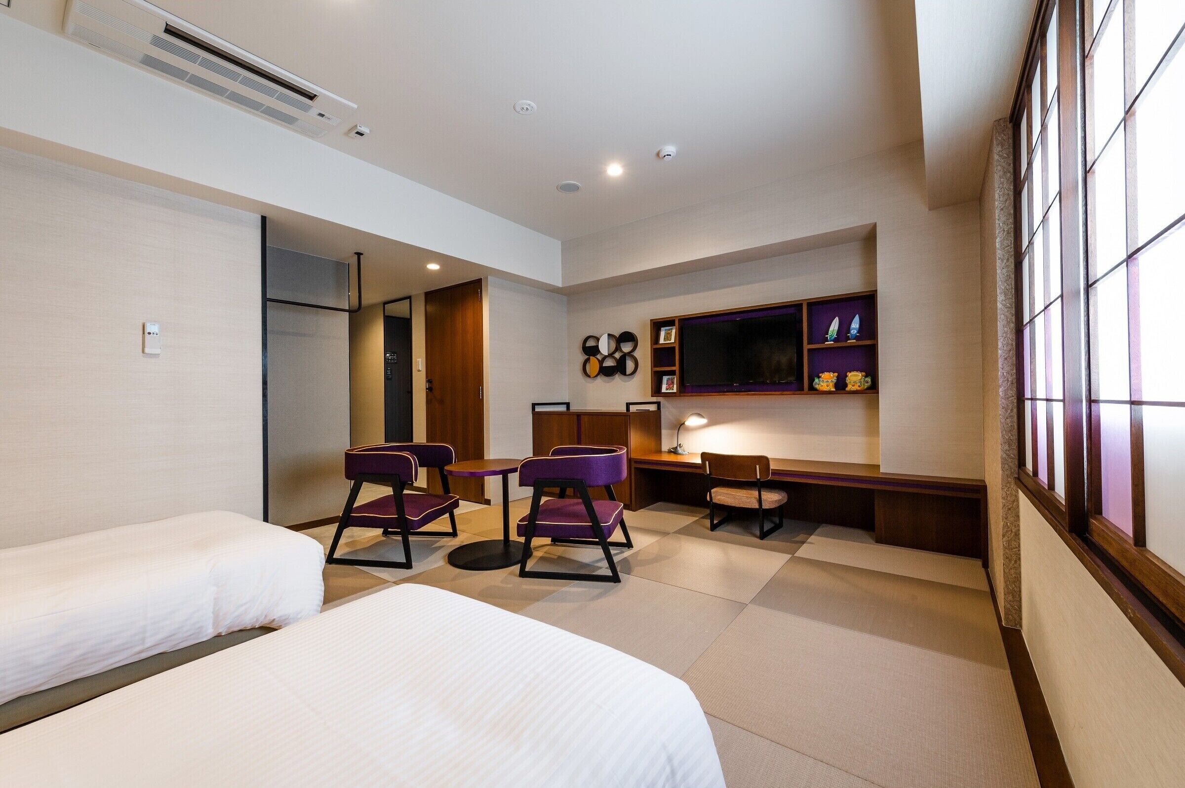 A hotel room with two beds, a desk, purple chairs, a TV, and a shelf with decorative items.