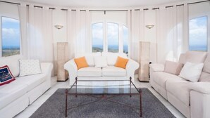 Un salon spacieux avec deux canapés blancs, une table basse en verre et de grandes fenêtres donnant sur le paysage.