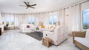 Un salon spacieux, meublé de meubles blancs, doté de grandes fenêtres et d’un ventilateur de plafond.