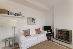 A white sofa with decorative pillows, a floor lamp, a bookshelf with books and a vase, a framed picture, a television, and a fireplace.