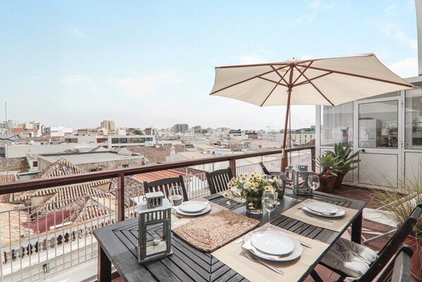 A rooftop terrace with a table set for dining, a large umbrella, and a view of the cityscape.