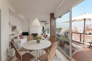 A modern living room with a white sofa, a dining table, wicker chairs, a fireplace, and a balcony with a glass door.