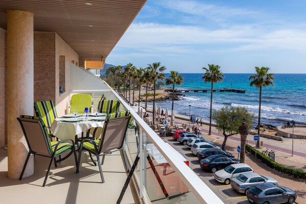 A balcony with a table and chairs, overlooking a beach with palm trees and parked cars.