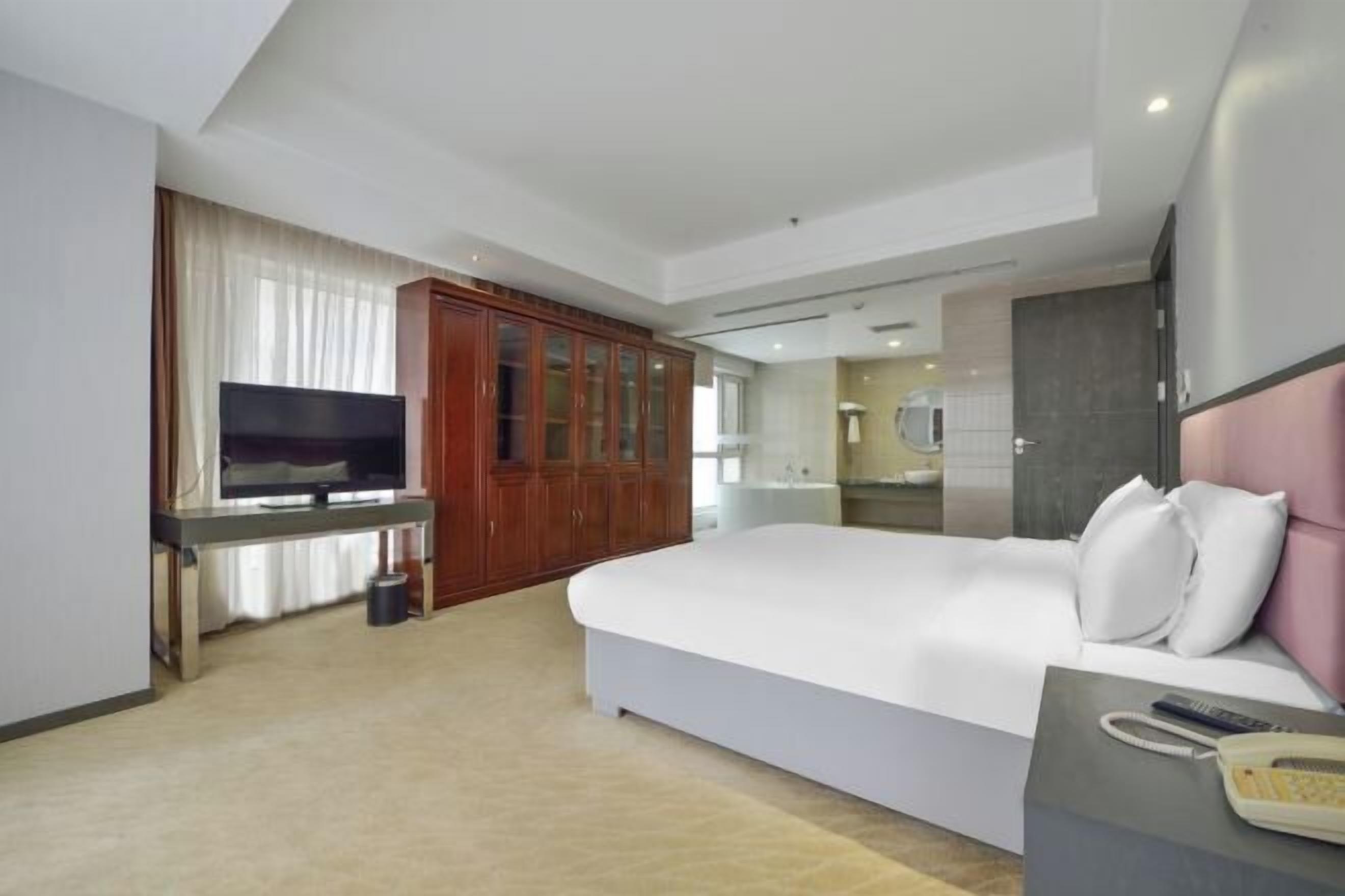 Ein modernes Hotelzimmer mit einem großen Bett, einem Fernseher, einem Holzschrank und einem durch eine offene Tür einsehbaren Badezimmer.