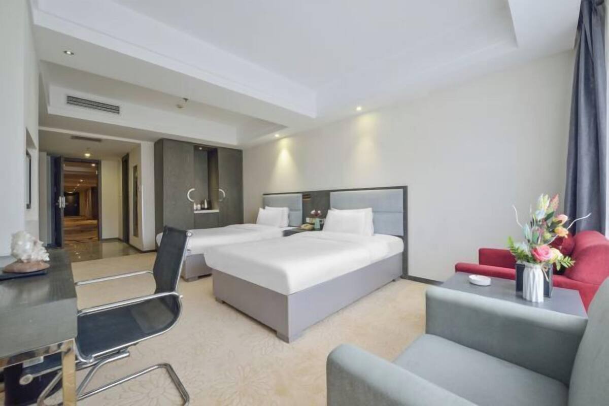 Une chambre d’hôtel avec deux lits, un bureau, une chaise, un canapé et une petite table sur laquelle est posé un vase de fleurs.