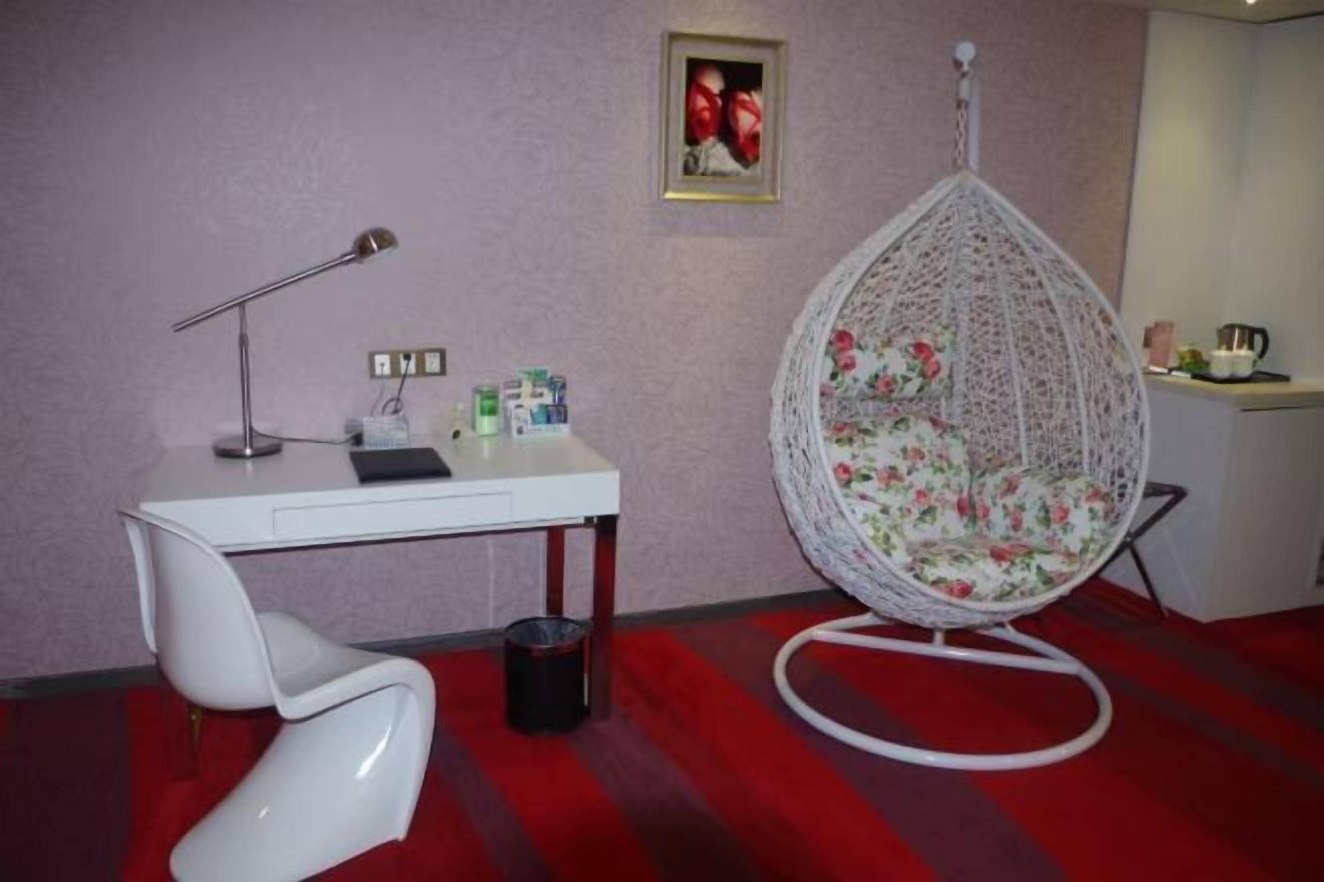 Une pièce avec un bureau blanc, une chaise blanche, un fauteuil suspendu recouvert d’un tissu à motifs floraux, un tableau encadré au mur et une moquette rouge.