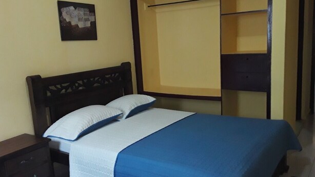 A neatly made bed with a blue bedspread and white pillows, a wooden headboard, a small bedside table, and a wall-mounted picture. - HABITACION DOBLE (Cartagena)