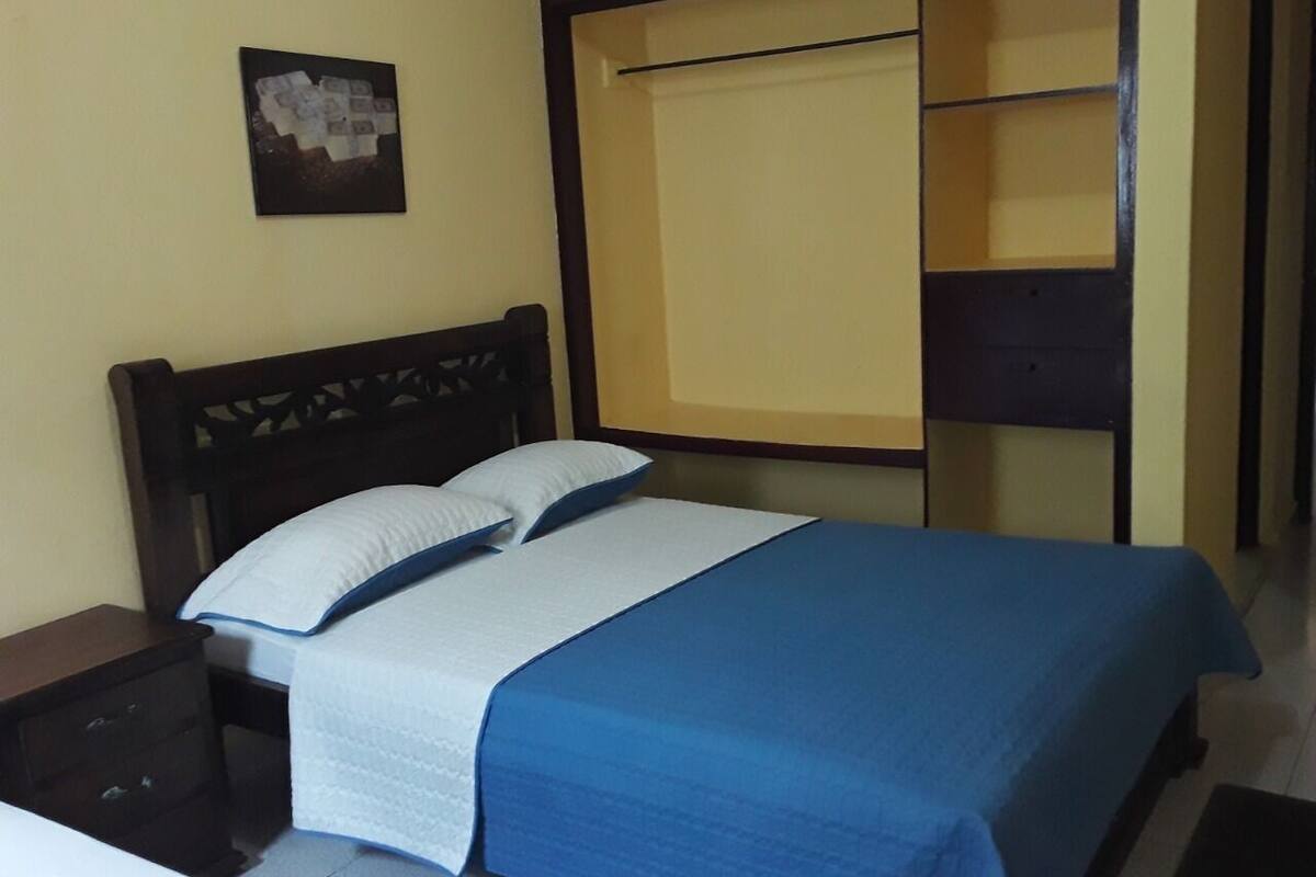 A neatly made bed with a blue bedspread and white pillows, a wooden headboard, a small bedside table, and a wall-mounted picture.