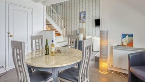 Un coin repas avec une table ronde, des chaises et une bouteille de vin. On aperçoit un escalier au fond, qui mène à une autre pièce.