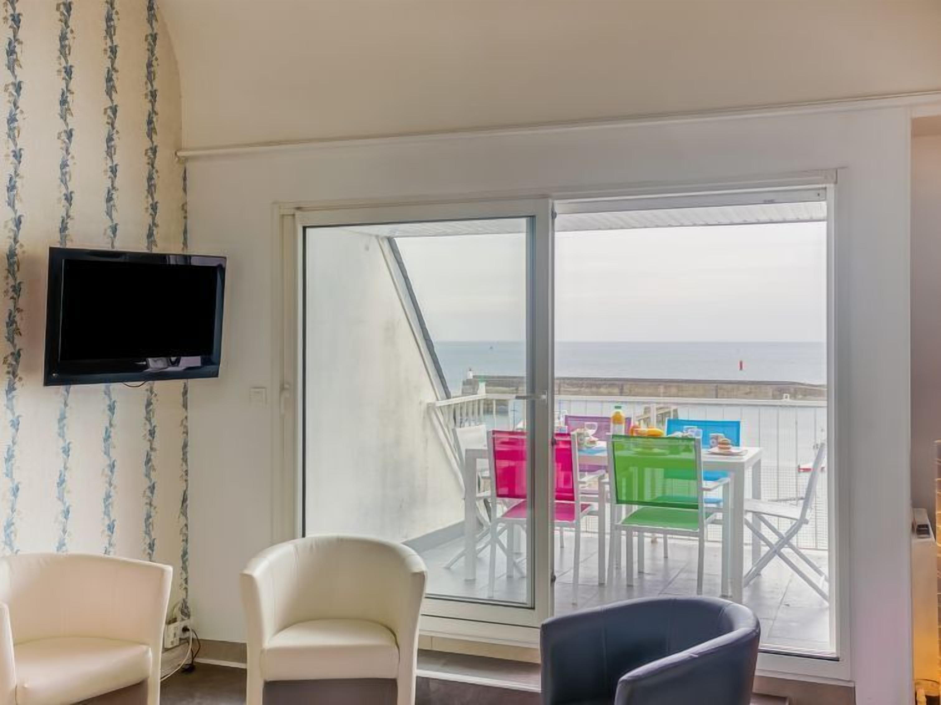 A room with a large sliding glass door leading to a balcony with colorful chairs and a table. A flat-screen TV is mounted on the wall.