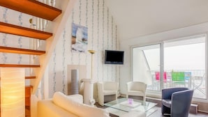 Un salon moderne avec un canapé, des fauteuils, une table basse en verre, un téléviseur et un balcon agrémenté de chaises colorées.