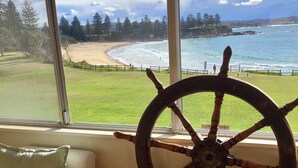 Een kamer met een groot raam dat uitkijkt op een strand en zee, een houten schippersrad decoratie, een groen en wit kussen, en een houten stoel.