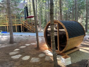 Una estructura de barriles de madera con una puerta, rodeada de árboles, y una terraza de madera con escaleras que conducen a un edificio.