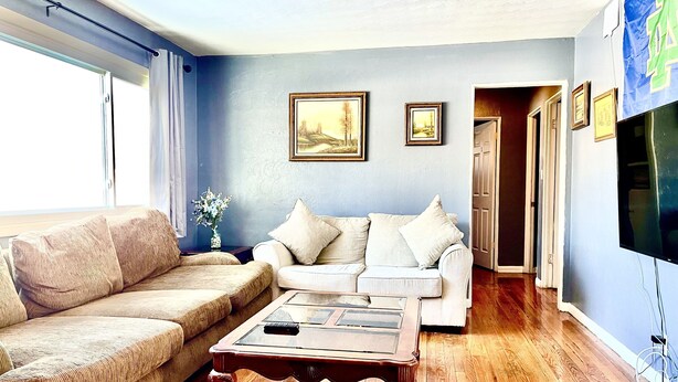 A living room with a beige sofa, a white sofa, and a wooden coffee table. There are two framed pictures on the wall, and a TV mounted on the wall.
