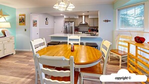 Uma sala de jantar com uma mesa de madeira redonda e cadeiras brancas, uma cozinha ao fundo e uma janela com persianas.
