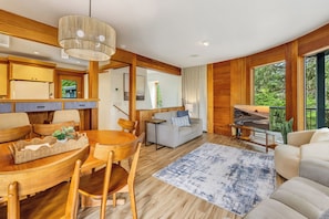 A modern interior with a wooden dining table, chairs, a sofa, and a large window offering a view of greenery.
