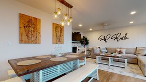 A modern living room with a wooden dining table, a sofa, and a coffee table. There are wall art pieces and a sign that reads "Surf Shack". - SURF SHACK at Seaside Beach- OCEAN FRONT - HOT TUB (Seaside)