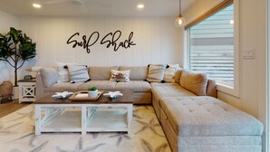 A living room with a sectional sofa, a coffee table, and a wall-mounted sign that reads "Surf Shack". - SURF SHACK at Seaside Beach- OCEAN FRONT - HOT TUB (Seaside)