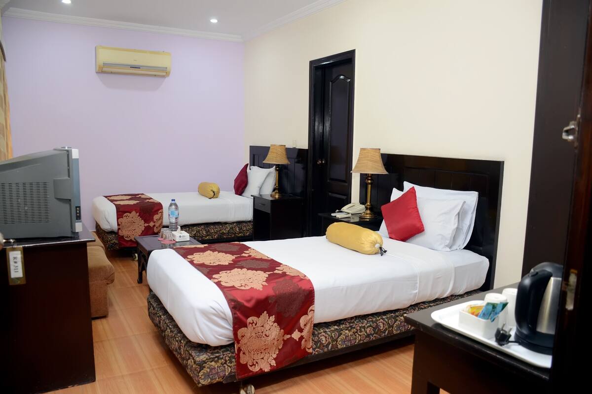 A hotel room with two beds, a TV, a desk with a kettle, and a bedside table with a lamp.