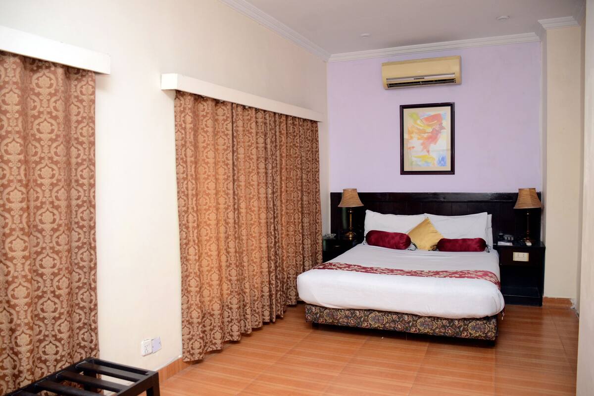A hotel room with a large bed, patterned curtains, an air conditioning unit, and a framed picture on the wall.