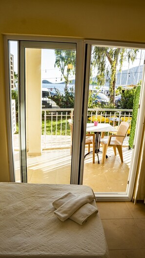 A room with a bed, a sliding glass door leading to an outdoor balcony with a table and chairs, and a view of the sea and buildings. - Kaju Rooms (Marmaris)