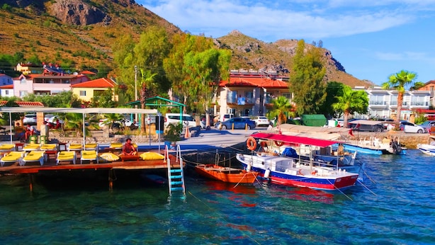 A marina with boats docked, a covered area with seating, and buildings in the background. - Kaju Rooms (Marmaris)