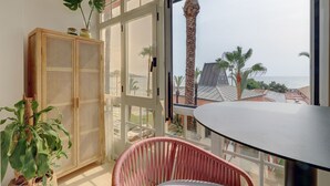 A balcony with a view of palm trees and a beach, a wicker cabinet, and a red chair. - Ale Maison sotogrande port and beachfront private terrace sea view (San Roque)