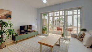 A living room with a TV, sofa, coffee table, and a large sliding glass door leading to an outdoor patio with chairs and plants. - Ale Maison sotogrande port and beachfront private terrace sea view (San Roque)