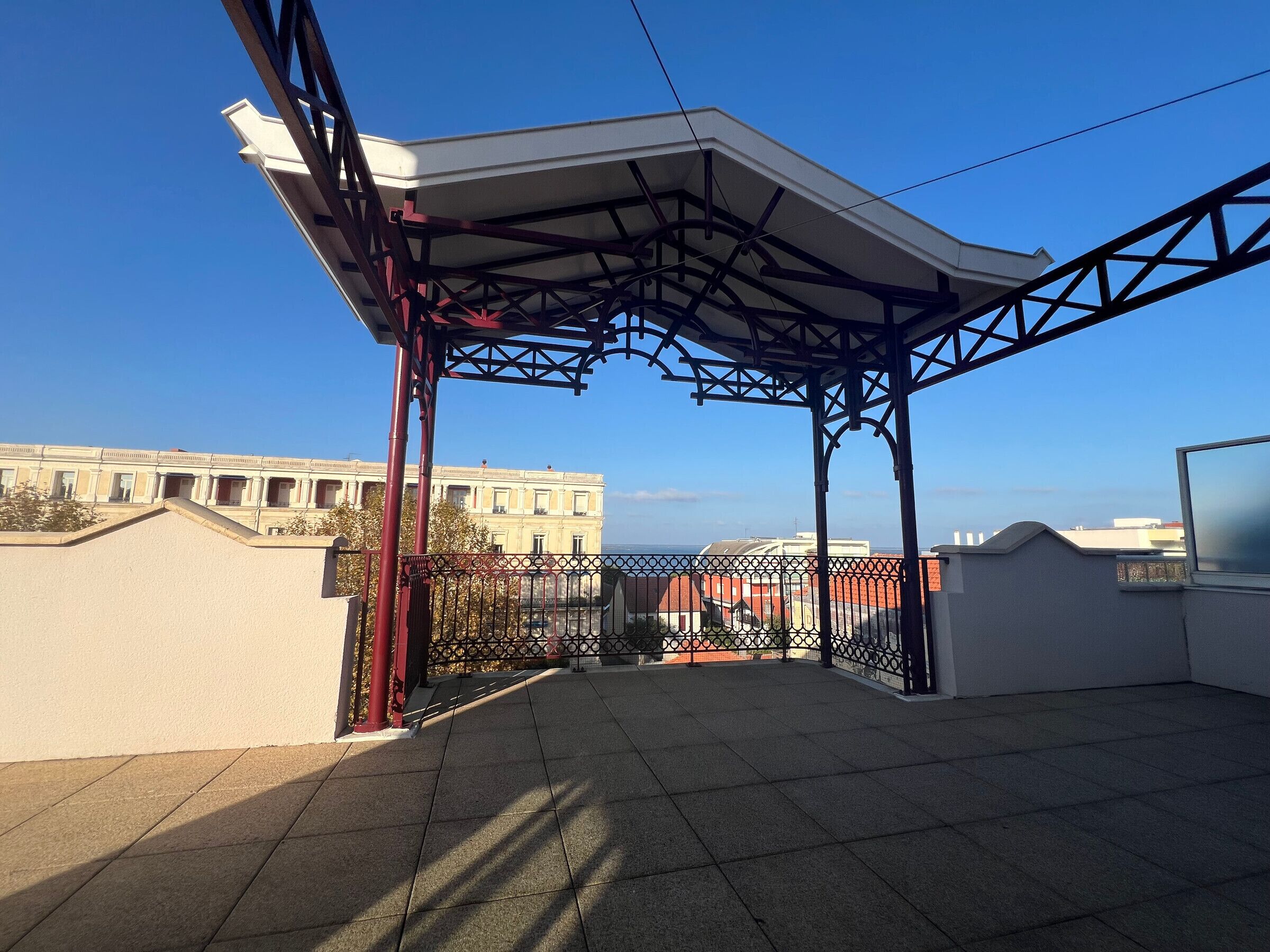 Ein überdachter Außenbereich mit Metallgeländer, Fliesenboden und Blick auf Gebäude im Hintergrund.