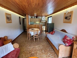 A cozy room with a wooden ceiling, a dining table with chairs, a sofa with patterned cushions, and a kitchen area in the background.