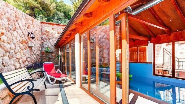 Zona junto a la piscina con pared de piedra, terraza de madera y puertas de cristal que dan a la piscina.