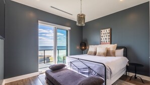 Un dormitorio con cama, una banca, una mesita de noche, una lámpara de pie y vista al mar.